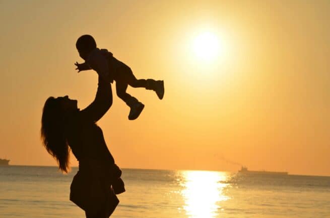 Bright sunset silhouette of a woman lifting a child by the beach with ships on the horizon.